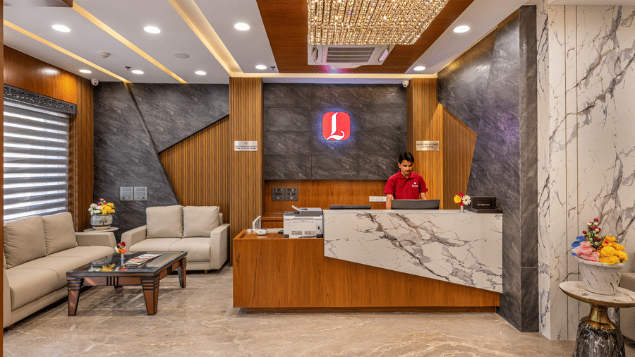 A staff member of Lords Hotels & Resorts, standing at reception desk of the best hotel in Sumerpur, Rajasthan, with a marble backdrop, exquisite ceiling details, and cosy lighting.
