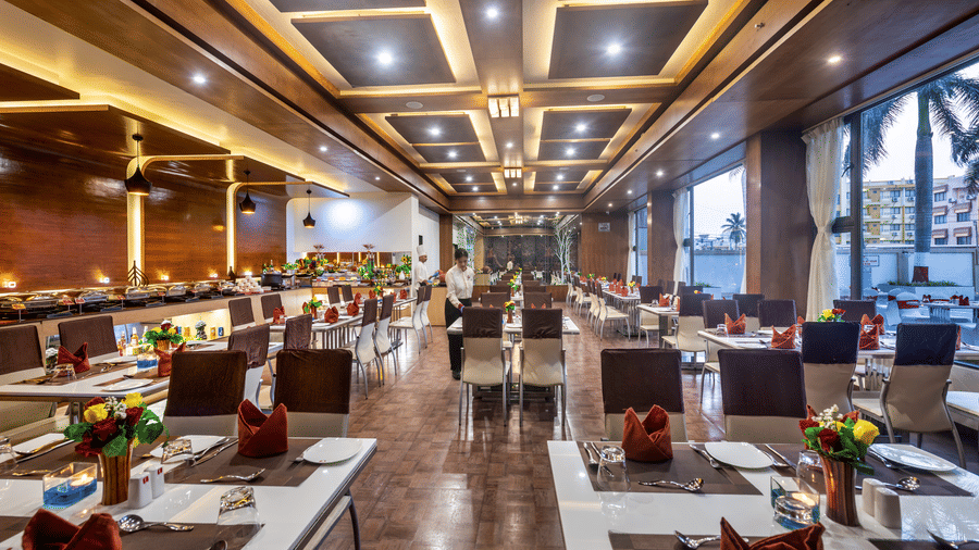 A spacious and elegantly designed restaurant with well-set tables adorned with flowers and large windows offering a view of the outdoors at Lords Inn Somnath.