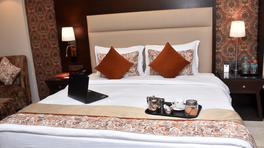 A hotel room at Lords Plaza Surat featuring a double bed decked with plush pillows, patterned runner, a large head-rest and a tray of cookies and tea on bed along with an open laptop