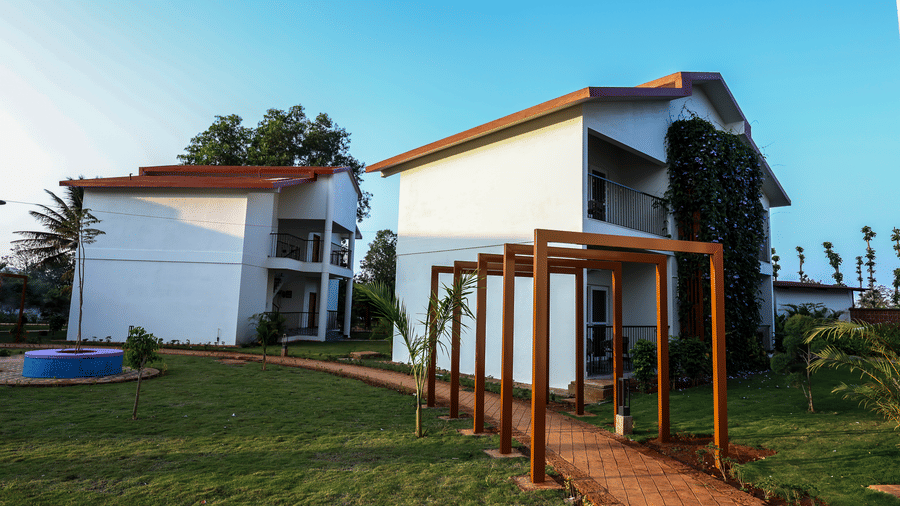 A wooden pergola walkway over a brick path leading across a green lawn toward white resort buildings | Lords Sahyadri Resort, Shivamogga