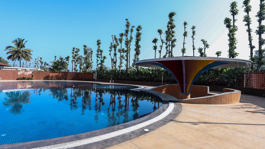 An outdoor swimming pool with blue water reflecting tall trees, next to a curved pool deck under a clear sky | Lords Sahyadri Resort, Shivamogga