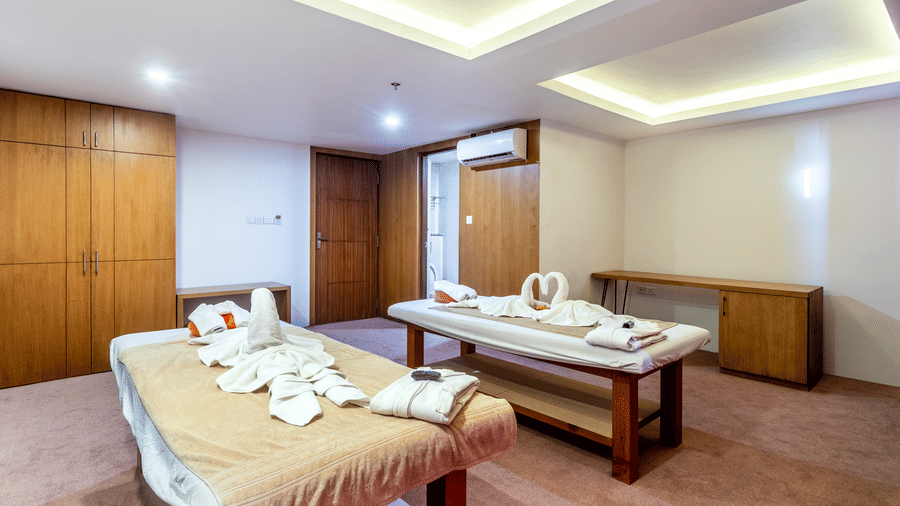 Turkey towels folded in swan-shape on spa tables in a room with AC, designed ceiling and a door opening to a restroom. 