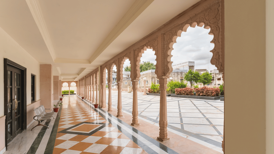 A covered outdoor corridor at Shri Vallabh Vilas Lords Plaza Nathdwara, with arched stone columns and checkered flooring, opening to a garden area.