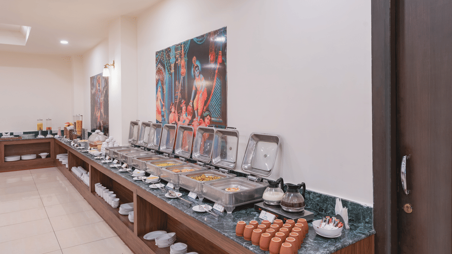 Buffet setup with covered serving dishes, beverages, and utensils arranged at the Manorath Restaurant at Shri Vallabh Vilas Lords Plaza Nathdwara, along a counter in a bright dining area.