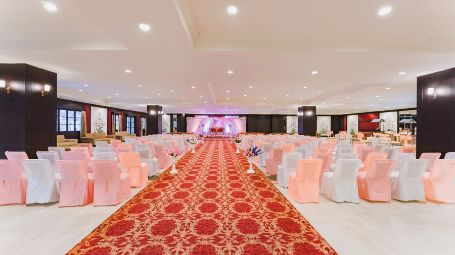A interior deccorated setup with a red carpet aisle leading to a stage, at the Raj Mahal at Shri Vallabh Vilas Lords Plaza Nathdwara, surrounded by neatly arranged peach chairs.