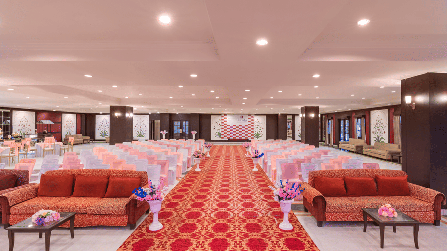 A wide red carpet aisle flanked by peach chairs and sofas in a large indoor Raj Mahal Hall at Shri Vallabh Vilas Lords Plaza Nathdwara.