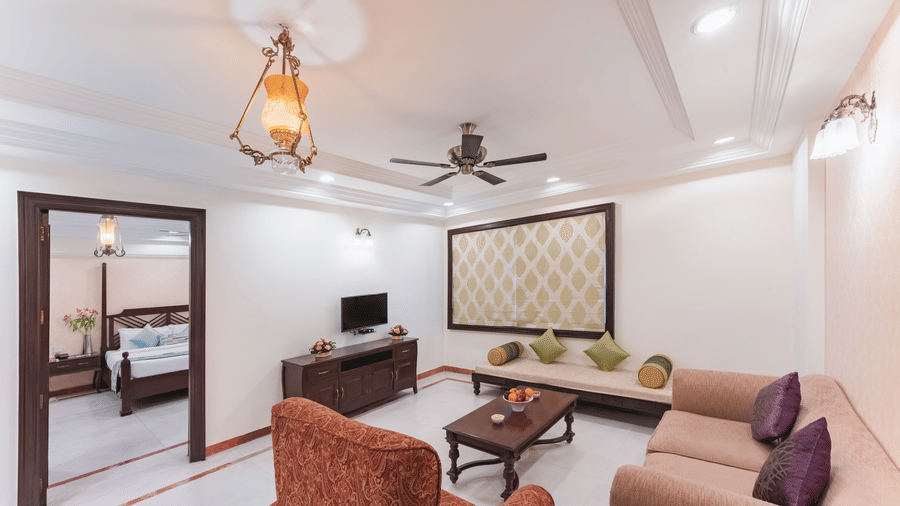 A view of a spacious living area in a deluxe suite at Shri Vallabh Vilas Lords Plaza Nathdwara, featuring pendant lights, fan, a patterned wall art and a T.V. 