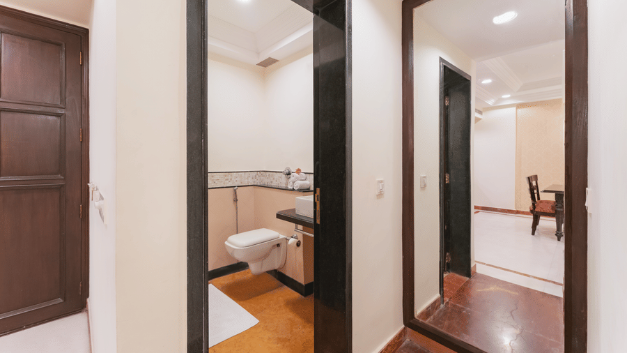 A well-lit corridor with polished flooring features a wall-mounted toilet in a compact bathroom on the left and a large full-length mirror on the right at Shri Vallabh Vilas Plaza Nathdwara.