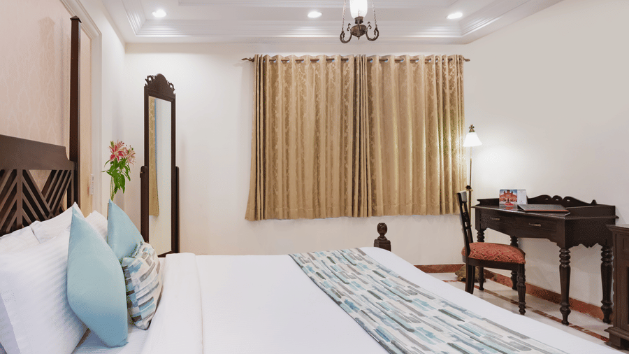 A view of a premier suite at Shri Vallabh Vilas Lords Plaza Nathdwara,featuring a twin size bed with full length mirror, wooden furnished table set, beige curtains, and pendant lights.