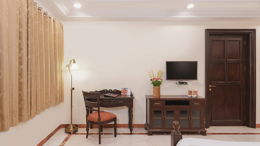 A cosy corner in the grand suite at Shri Vallabh Vilas Lords Plaza Nathdwara featuring a writing desk, floor lamp, television, and elegant beige curtains.