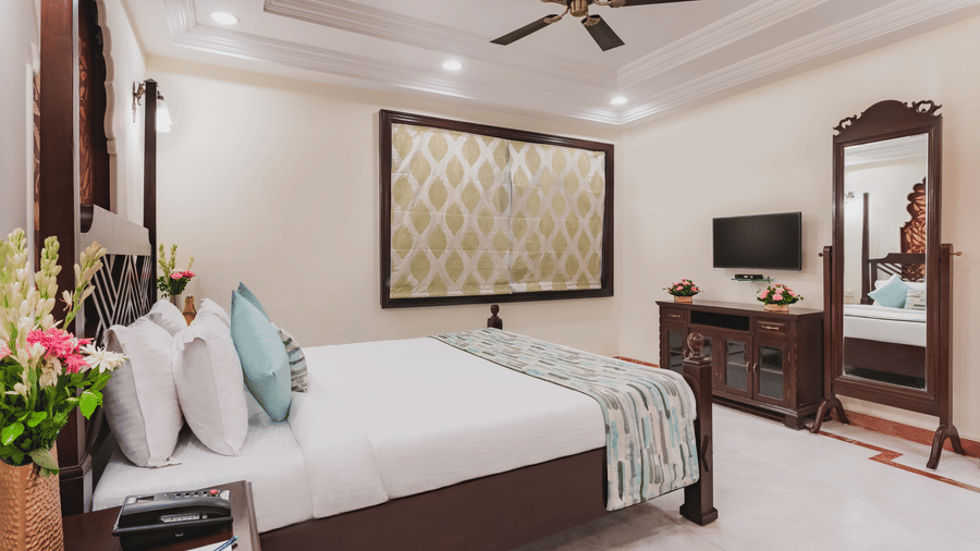 A side angle view of an elegant room at Shri Vallabh Vilas Lords Plaza Nathdwara, featuring twin size bed, be, wooden furnishings, wall art, a full length mirror and a T.V. above a cabinet.