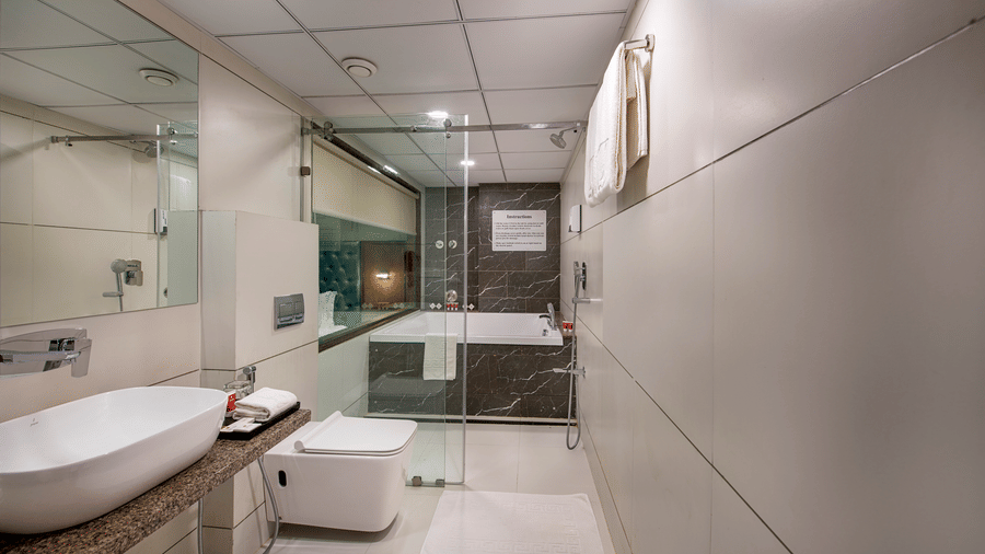 A modern hotel bathroom in Bengaluru features a sleek white sink on a stone countertop, a toilet, and a glass-enclosed bathtub - SK Lords Inn Ahmedabad