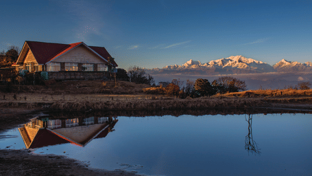 A building with a red roof overlooks a pond reflecting its structure, with mountains in the background.