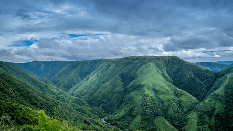Aerial view of a lush green mountain valley with a river winding through the base, under a partly cloudy blue sky.