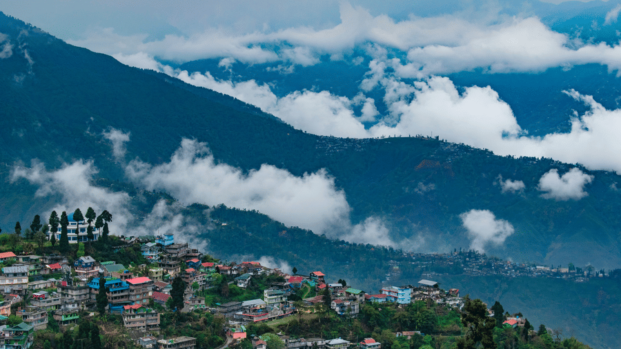 A hillside town nestled on a mountain slope with colorful buildings, dense trees, and misty clouds above.