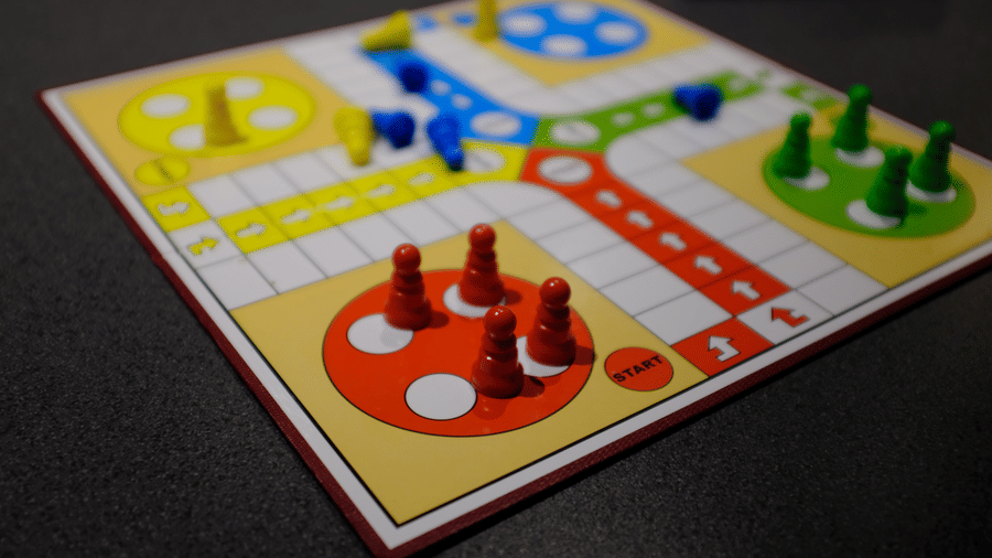 Close-up of a Ludo board game with red, yellow, blue, and green pieces resting on their coloured starting spots against a dark background.