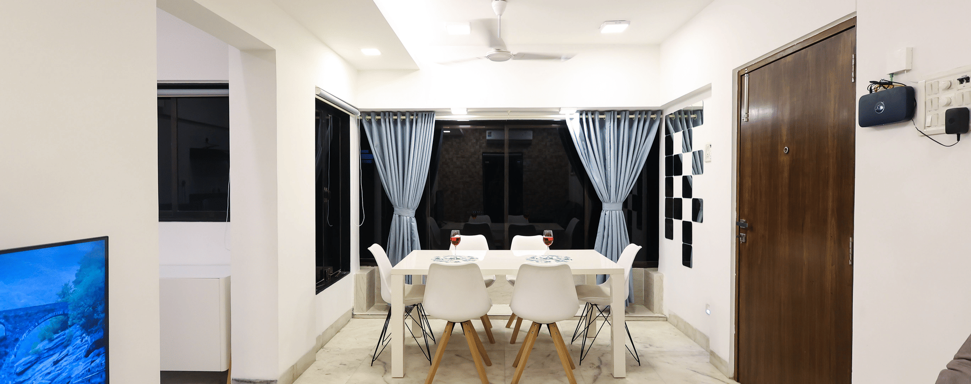 Dining area at Wabi-Sabi, Juhu with a white 6 seater table, moulded chairs, blue grey curtains, a ceiling fan, and a dark wooden door.