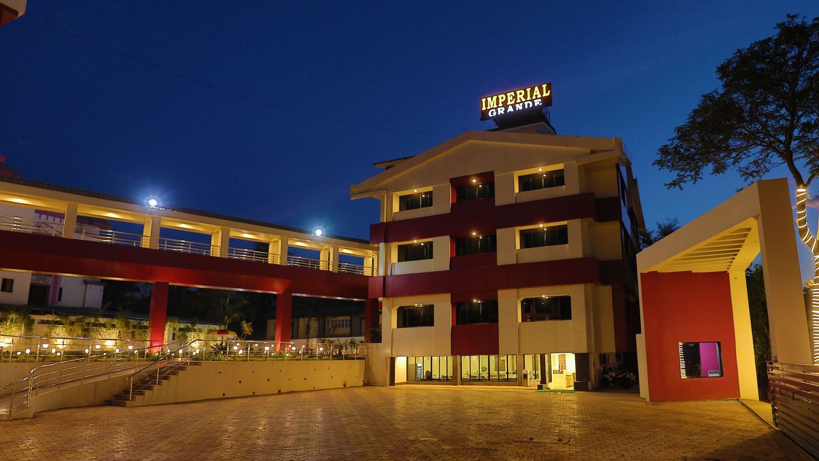 The exterior of Imperial Grande at night, showing a multi-story building with illuminated logo and a paved courtyard reflecting the lights.