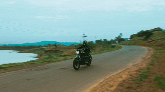A biker driving down an narrow empty street on his bike with the road having a small water body on one side and some grassland on the other.