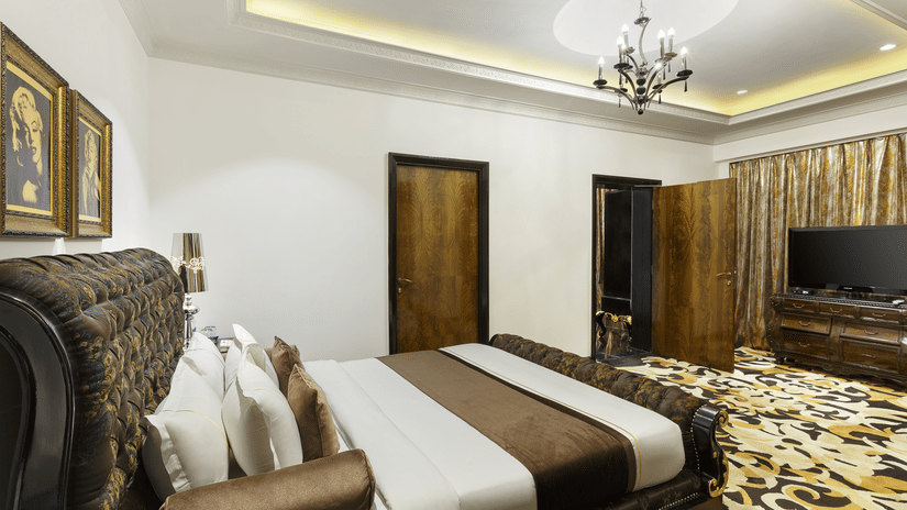 The spacious presidential suite bedroom with a king size bed, upholstered headboard, bedside tables, chandelier lighting, television unit and patterned flooring at La Marvella, Bengaluru.