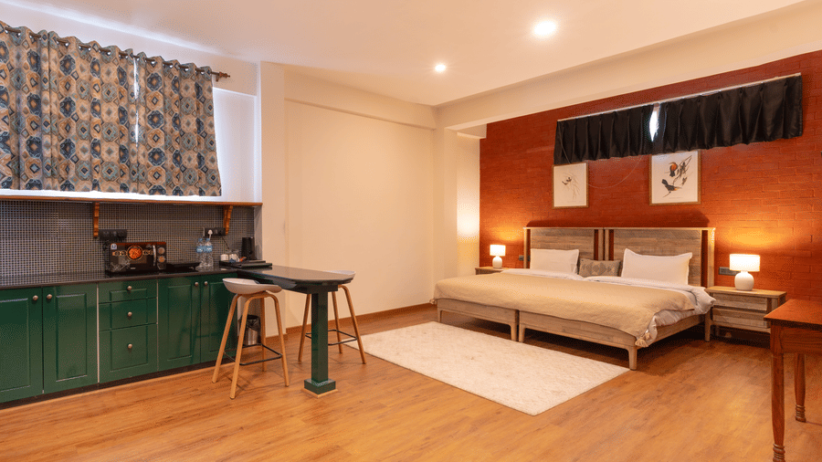 An open-plan studio apartment showing the green cabinetry of the kitchenette and the sleeping area with warm lighting - Johnson Lodge & Spa, Manali