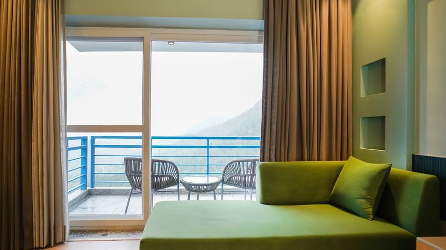 A modern hotel room with a green sofa and a large sliding glass door opening to a balcony with outdoor seating and a view of the hills | Deluxe Room with Valley View | Sukama Resort & Spa, Nainital
