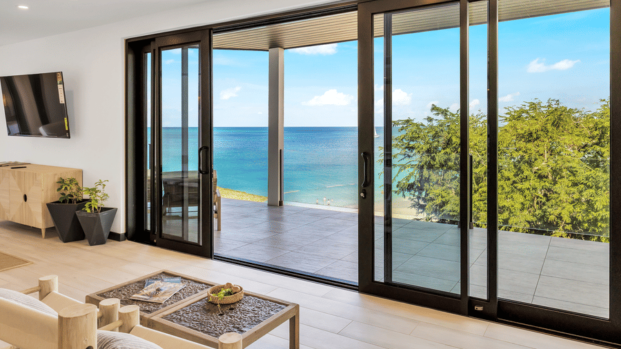 A living area with a lounge chair, a table, a wall mounted television, a drawer cabinet, and a large glass sliding door leading to a patio with an ocean view at Tamarind Hills Resort and Villas, Antigua.