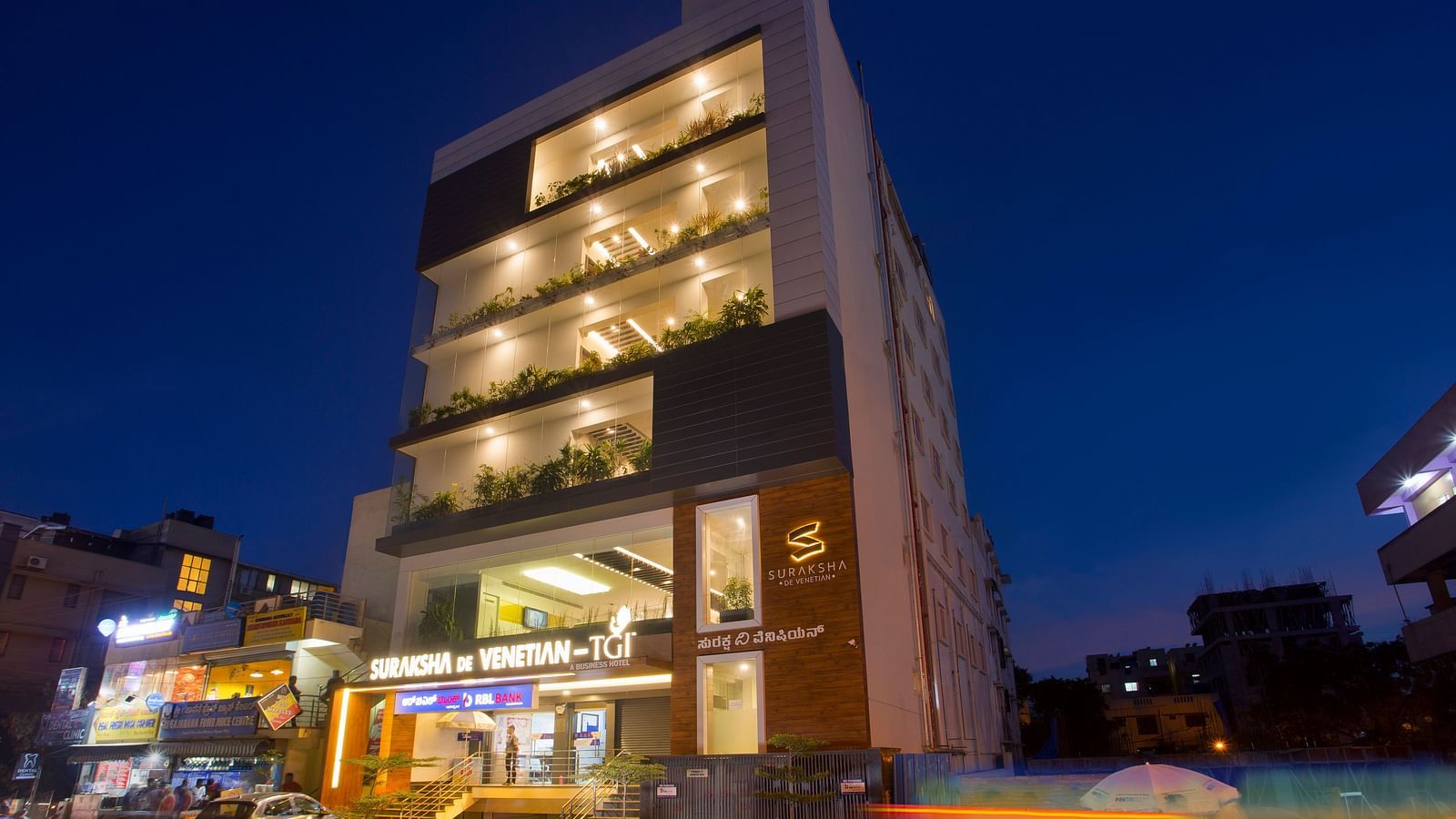 An exterior view of a modern multi-story building illuminated at dusk with signage and street activity - De Venetian by TGI, Brookefield