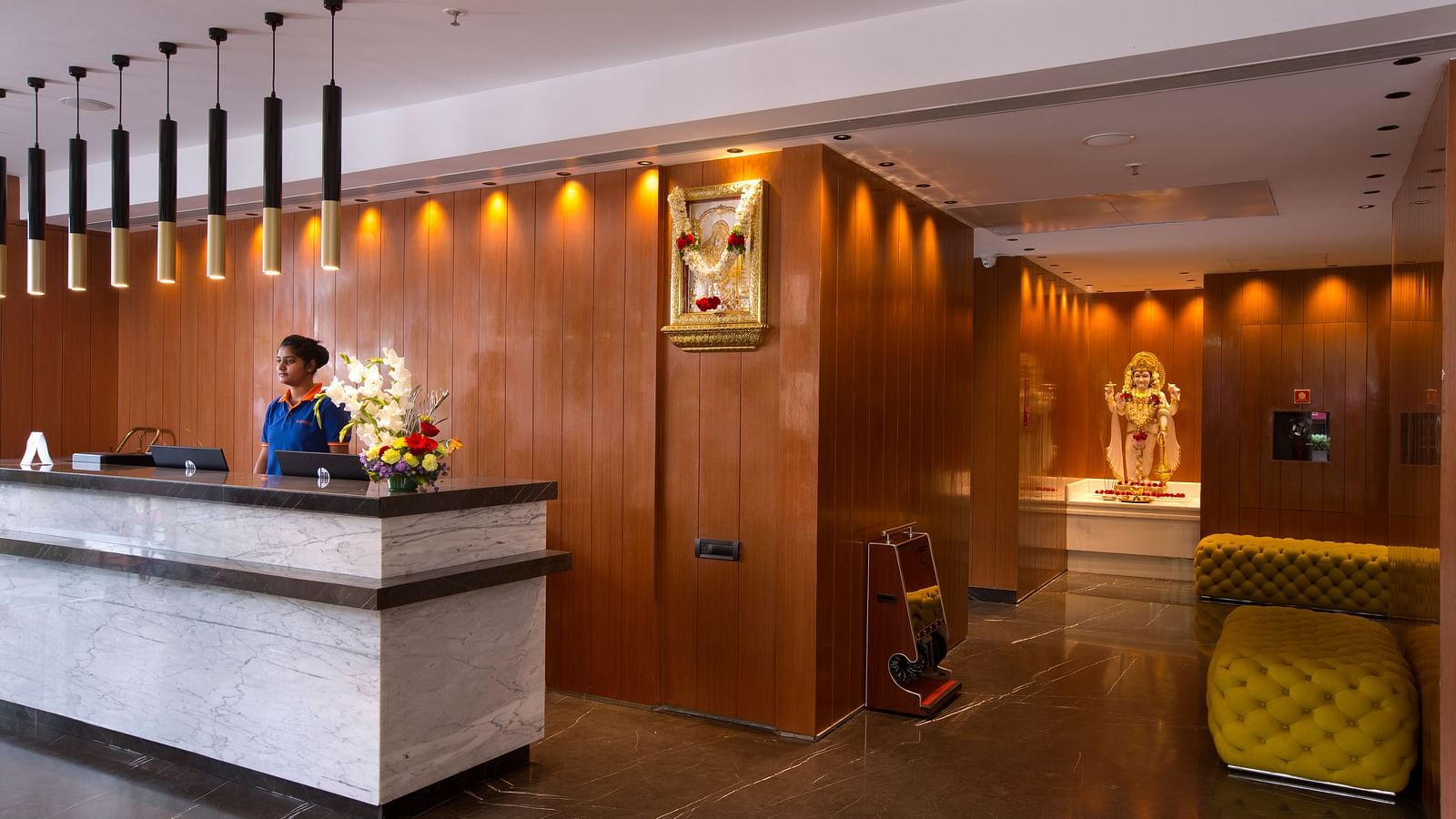 The hotel reception desk with a wooden backdrop and a flower arrangement, flanked by a statue of god in a glass case - Hotel Southend by TGI - Bommasandra, Bangalore