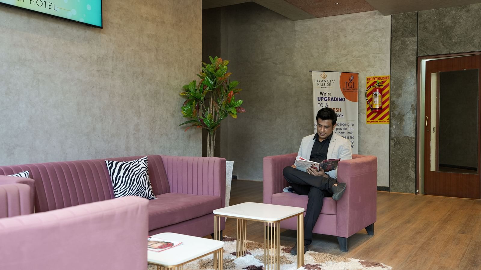 Man seated in the lounge area reading a magazine, surrounded by pink seating and indoor plants at TGI Livancia Hillside, JC Nagar, Mysore