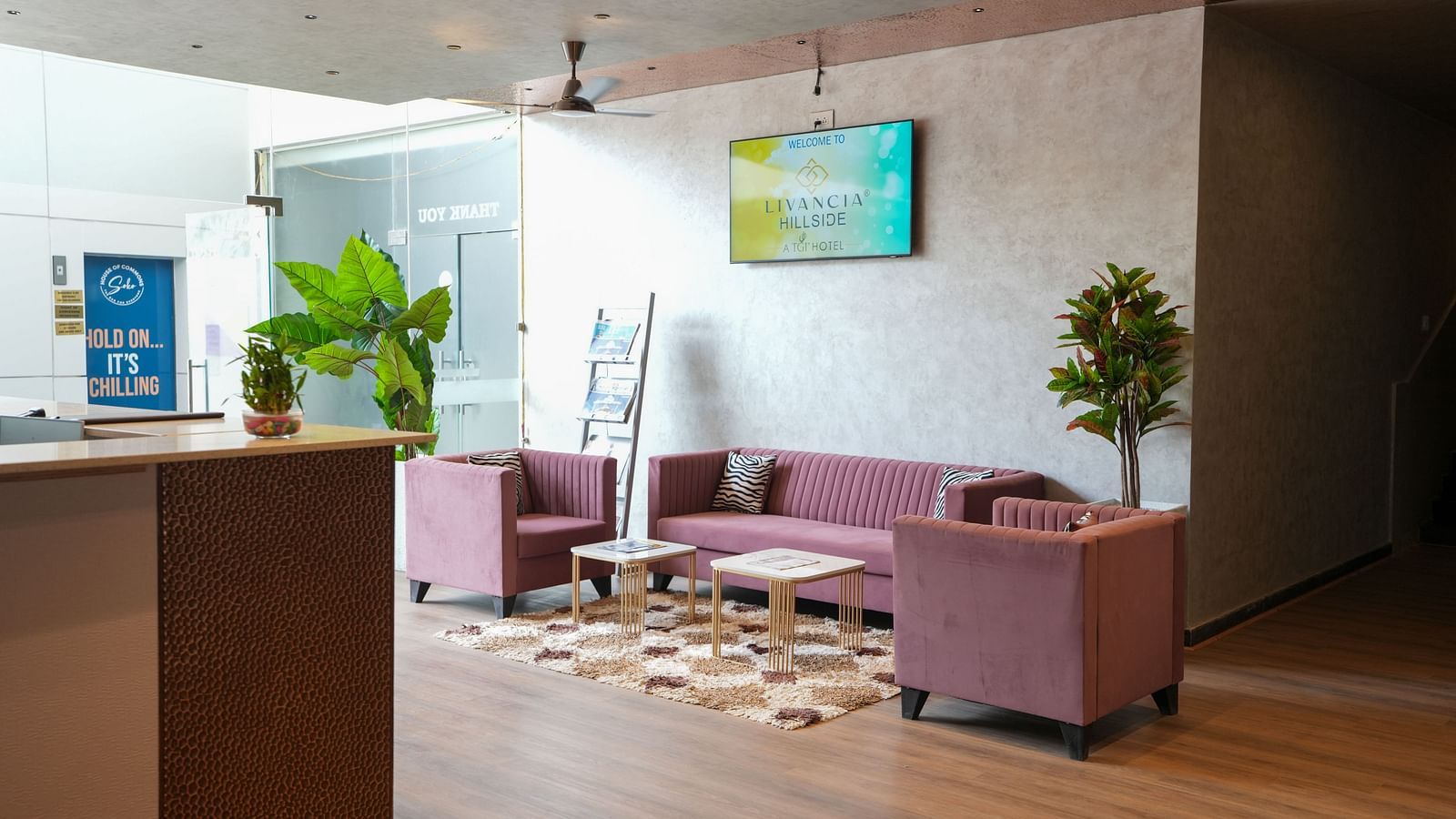 Lobby seating with pink sofas and a patterned rug near glass doors and indoor plants at TGI Livancia Hillside, JC Nagar, Mysore