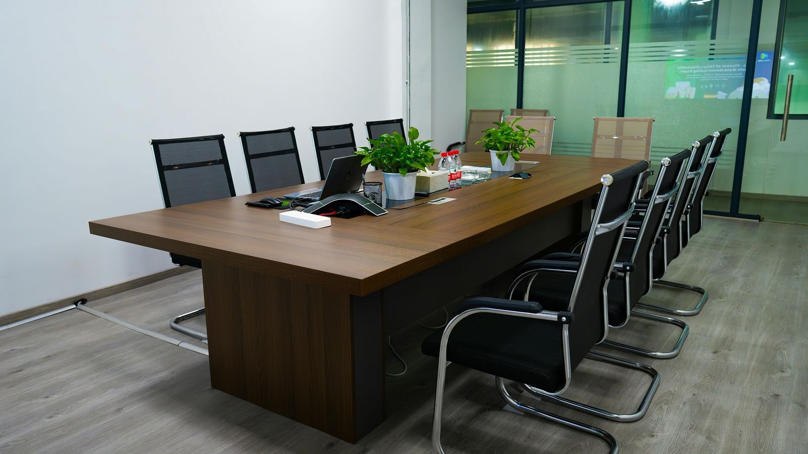A modern conference room with a long wooden table and black office chairs, under bright lighting.