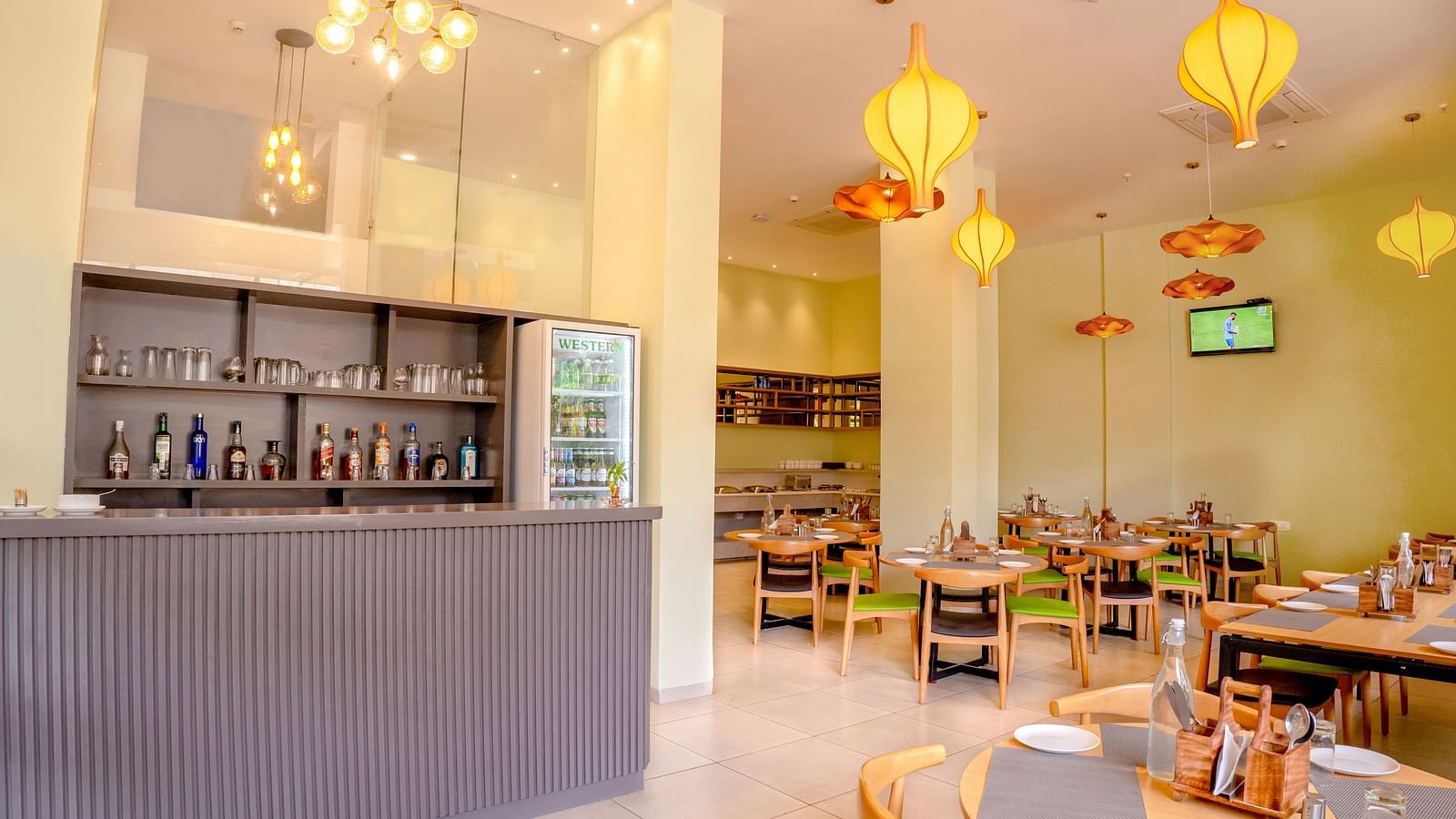 A spacious restaurant with a bar and beverage fridge on the left and dining tables with orange and green chairs under hanging orange lights - TGI Ameya Grand, Goa