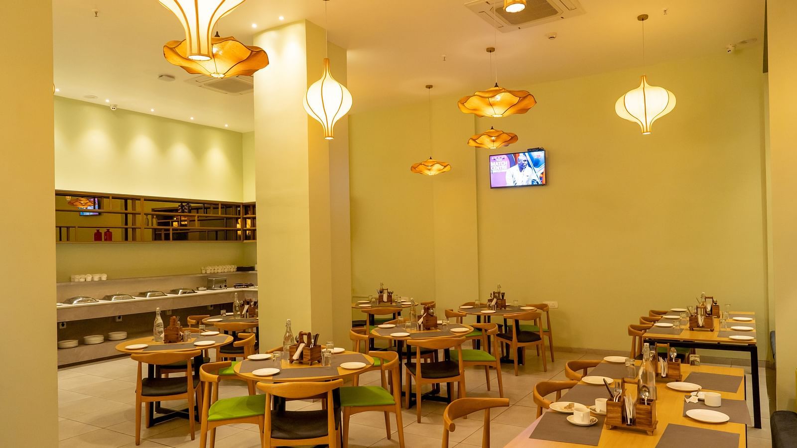 Another angle of the restaurant interior showing tables set for dining with hanging orange pendant lights and a TV screen on the wall - TGI Ameya Grand, Goa