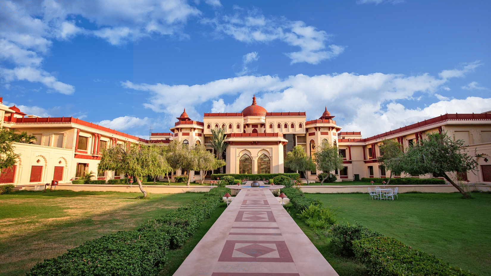 Grand exterior view of The Ummed Jodhpur Palace Resort & Spa with a pathway, lush lawns, and sky.