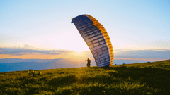 Colourful paraglider preparing for take-off across grassy hills, silhouetted against a warm sunrise with expansive open countryside beyond