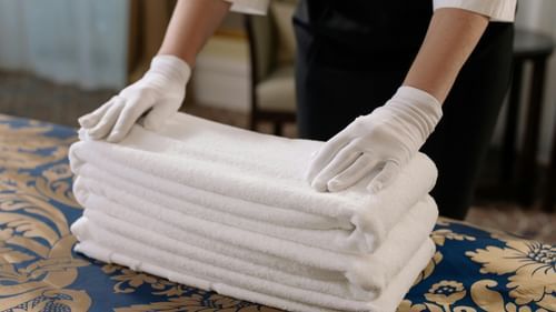 A hotel worker wearing white gloves neatly stacks fresh white towels on a patterned bedspread in a room.