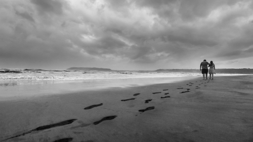 An image of a couple walking hand-in-hand on the shores of a calm beach leaving their footprints behind on the sand