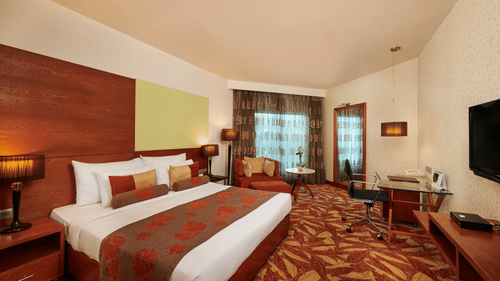 Room at Pride Premier Nagpur featuring a plush bed with pillows and cushions placed in front of a TV and a sitting space next to the window