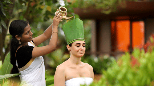 A woman in traditional attire pours herbal oil onto a relaxed guest’s head, covered with a green leaf wrap, in a serene setting at Uday Backwater Resort