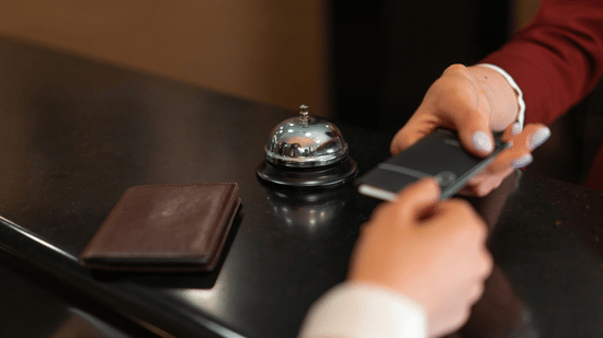 A close-up shot of a hotel receptionist in a red uniform handing a key card to a guest over a dark counter with a service bell.
