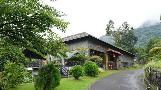 A charming wooden cottage-style building with its exterior lights on, situated on a green lawn with tall trees, under an overcast sky. | The Wild Trails Munnar