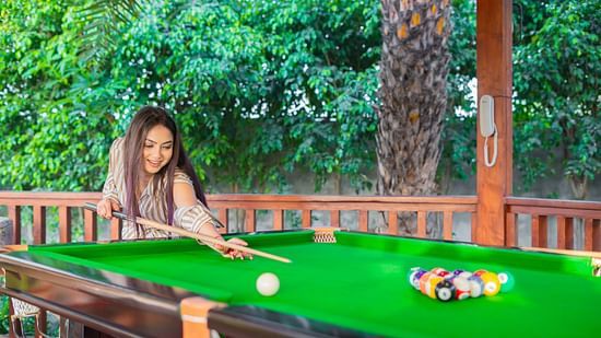 A woman with long dark hair playing pool on a green table in a well-lit outdoor or semi-outdoor setting with lush greenery at Themis Farm House