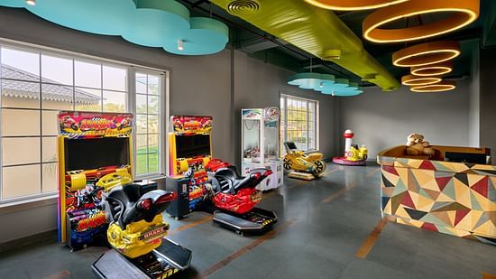 Vibrant indoor game room with colourful ceiling designs, arcade racing games, kiddie rides, claw machine, and a patterned reception counter, illuminated by large windows and circular ceiling lights - Voyage The Solitaire Bliss.