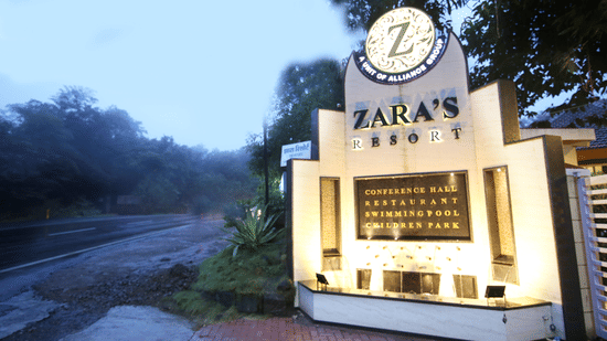 Entrance signage of Zara's Resort, Khandala, illuminated at dusk with surrounding greenery and a misty roadside setting.