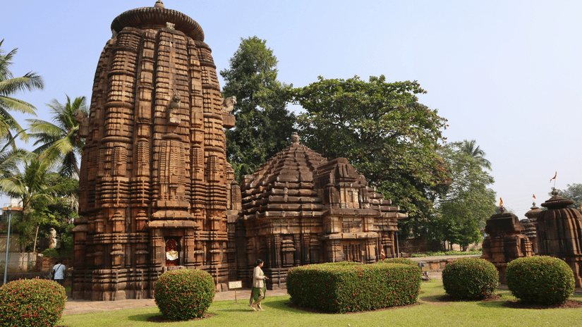 Ancient stone Mukteswar Temple, Bhubaneswar complex with carved towers, and a flag, surrounded by trees.