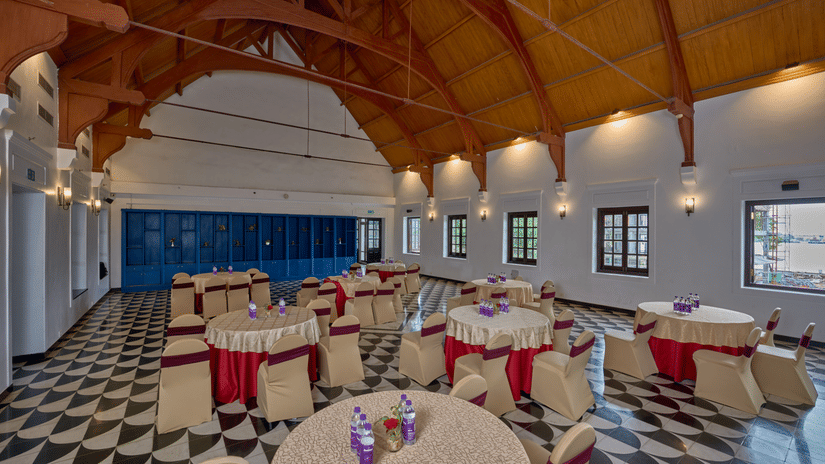 Spacious banquet hall at, The Old Court House by Abad, a business hotel in Kochi, featuring round tables, chequered floors, and an open wooden truss ceiling.
