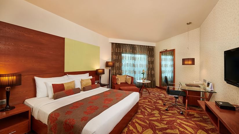 Room at Pride Hotel Nagpur featuring a plush bed with pillows and cushions placed in front of a TV and a sitting space next to the window