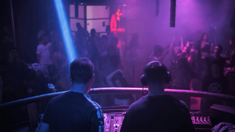 2 DJs performing at a console with stage lights, smoke effects, and a crowd visible in front of them.
