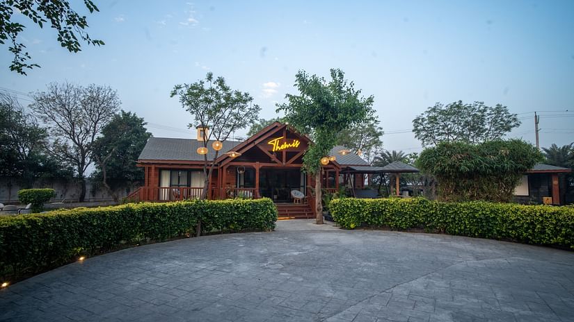 Front view of Themis Farm House with warm lighting, wooden exterior, lush greenery, and a spacious driveway under a clear evening sky.